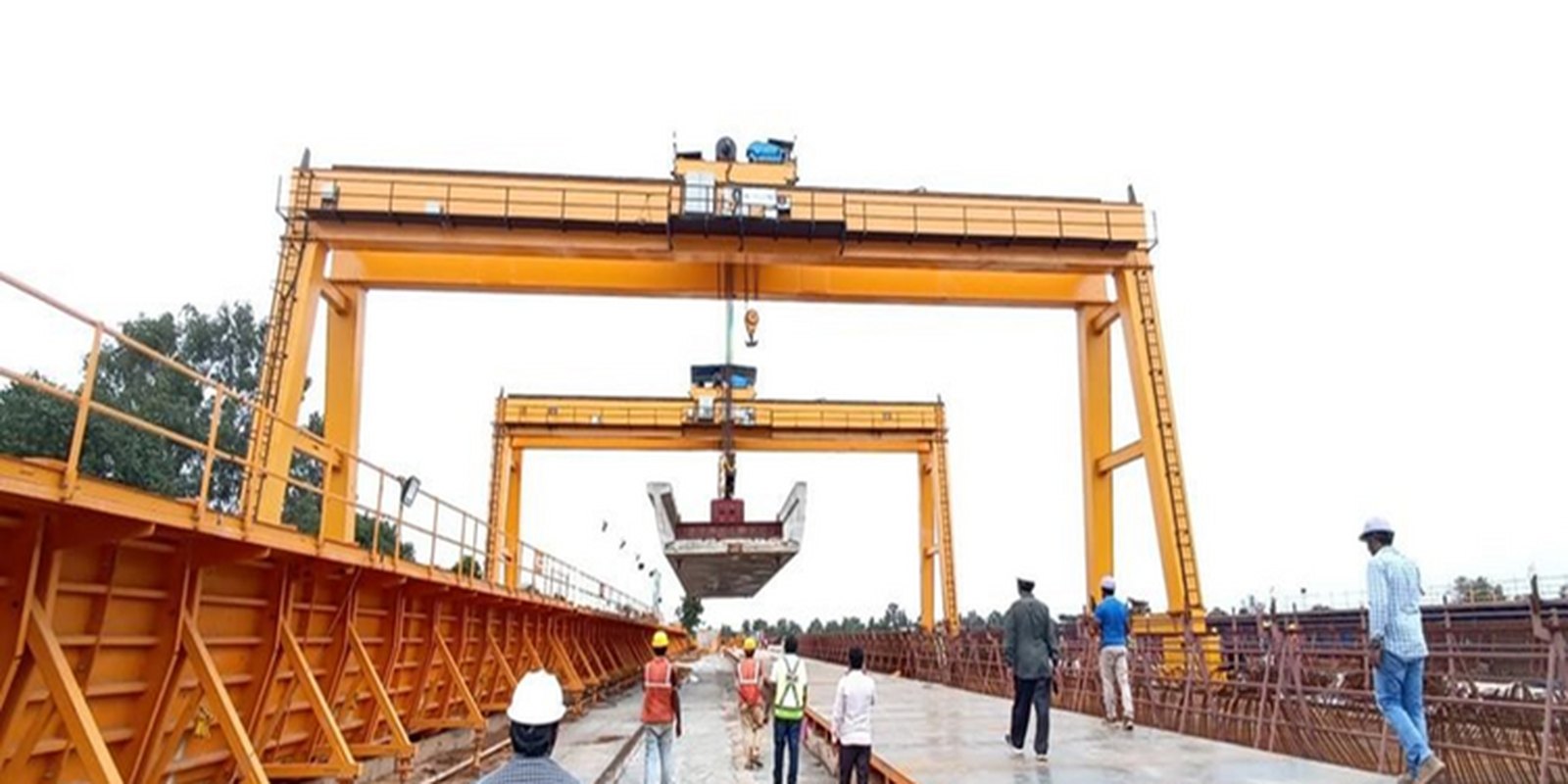 Double-girder goliath crane lifting a heavy segment mould while workers in high-visibility jackets guide the load